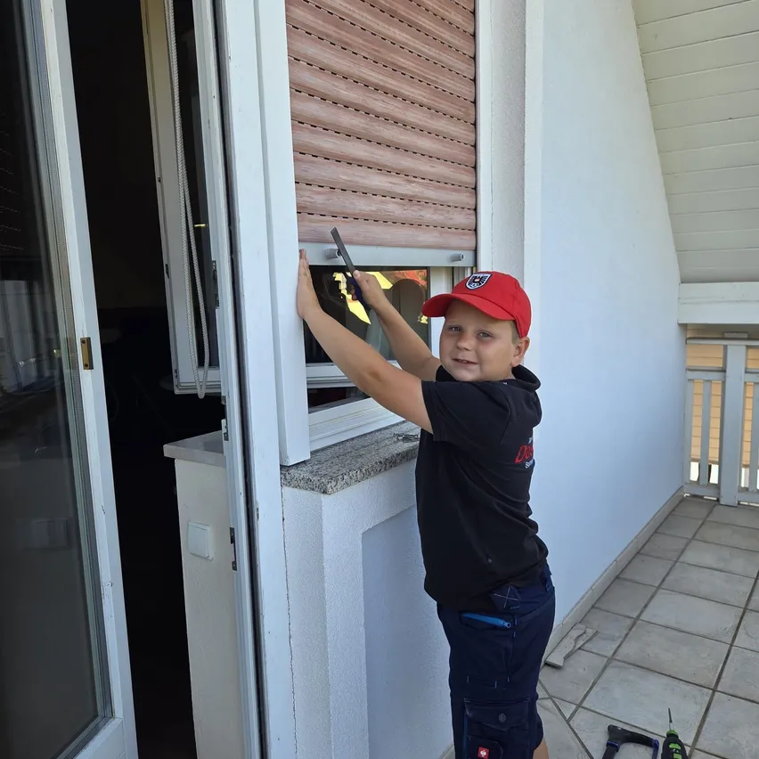 Ein junger Junge in einer roten Kappe und einem schwarzen Shirt repariert ein Fenster auf einem Balkon und hält ein Werkzeug. Der Balkon hat geflieste Böden und weiße Geländer.