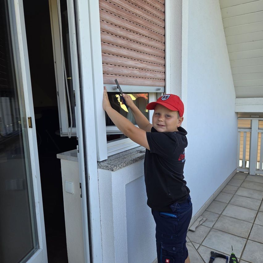 Ein junger Junge in einer roten Kappe und einem schwarzen Shirt repariert ein Fenster auf einem Balkon und hält ein Werkzeug. Der Balkon hat geflieste Böden und weiße Geländer.