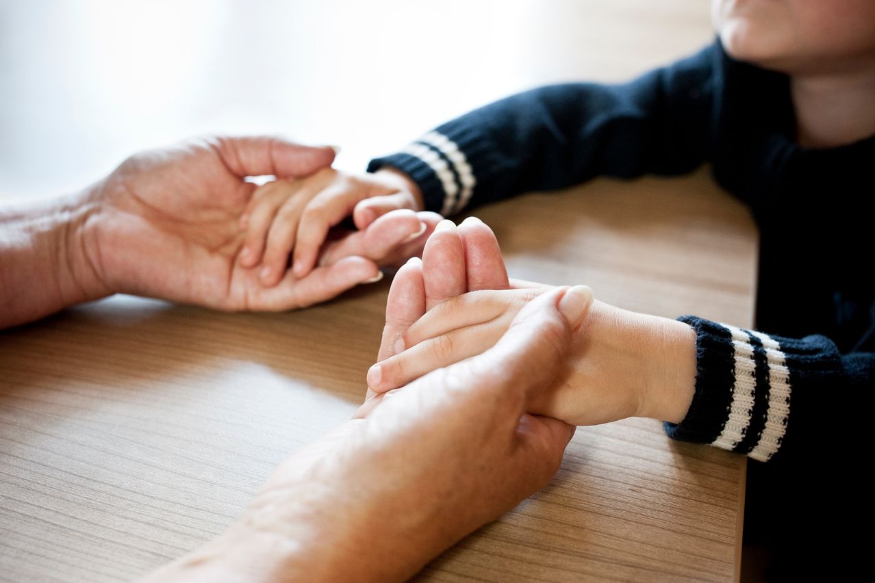 Zwei Hände, eine eines Erwachsenen und eine eines Kindes, halten sich auf einem Holztisch. Die Hand des Kindes hat weißen Nagellack.
