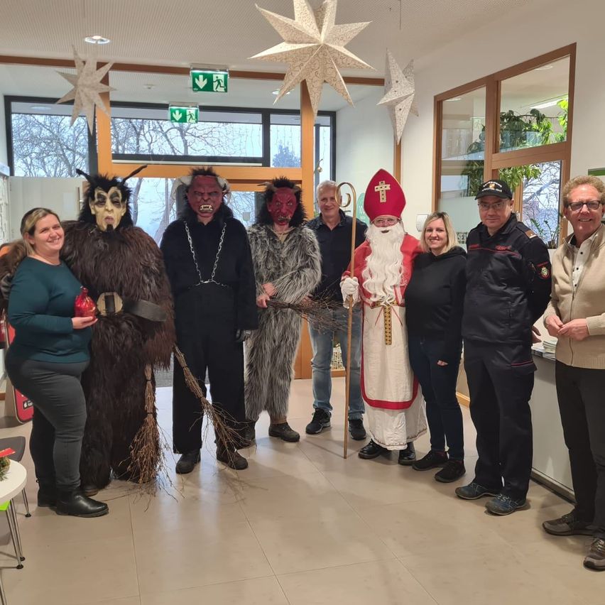 Eine Gruppe von Menschen posiert für ein Foto, einige in Kostümen, darunter ein Weihnachtsmann, eine Hexe und ein Teufel. Sie stehen in einem Raum mit gefliestem Boden, dekorativen Sternen und Glasfenstern.