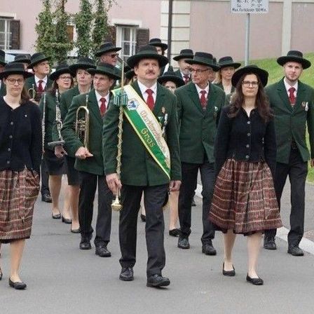 Eine Gruppe von Menschen in grünen Uniformen und Hüten marschiert in einem Umzug und trägt Musikinstrumente. Ein Mann mit einem Band steht in der Mitte.
