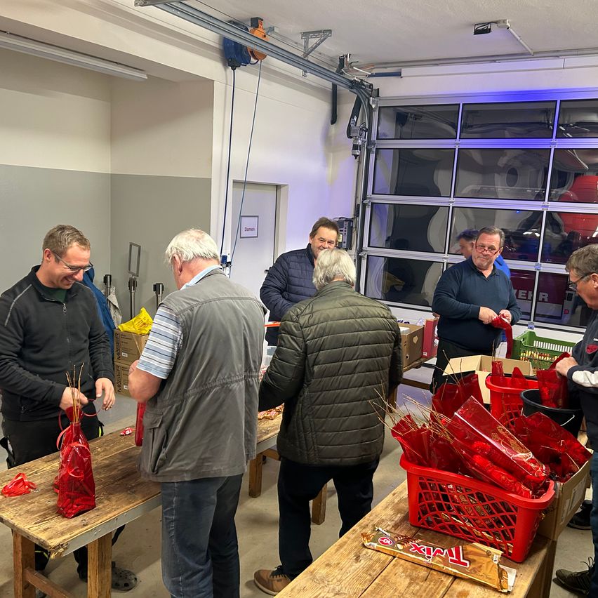 Mehrere Männer sind um einen Tisch versammelt und verpacken Geschenke in rote Tüten. Ein Mann trägt eine Brille und eine Jacke, ein anderer hat weiße Haare. Sie befinden sich in einem Raum mit einem großen Fenster und einem Schild an der Wand.