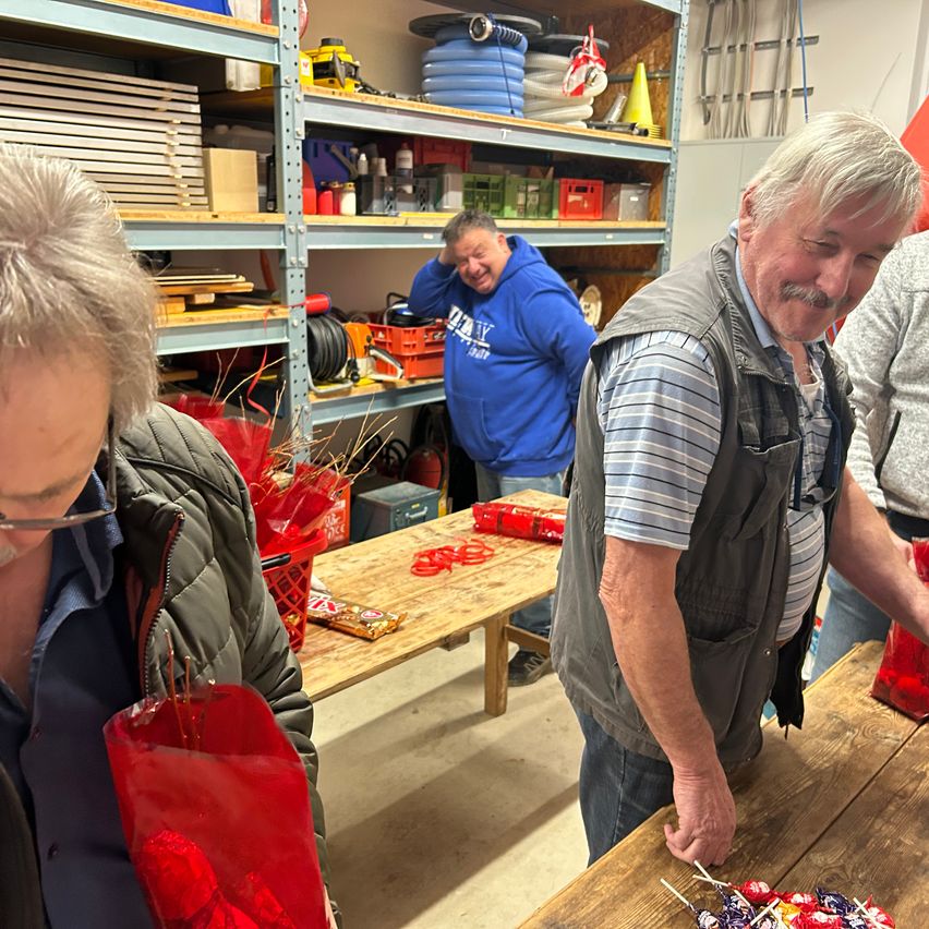 Eine Gruppe von Menschen befindet sich in einem Raum mit Regalen, die mit verschiedenen Gegenständen gefüllt sind. Ein Mann in einer Weste lächelt und hält eine Tüte mit Süßigkeiten. Ein anderer Mann in einer Kapuzenjacke steht hinter ihm. Eine Frau hält eine rote Tasche.