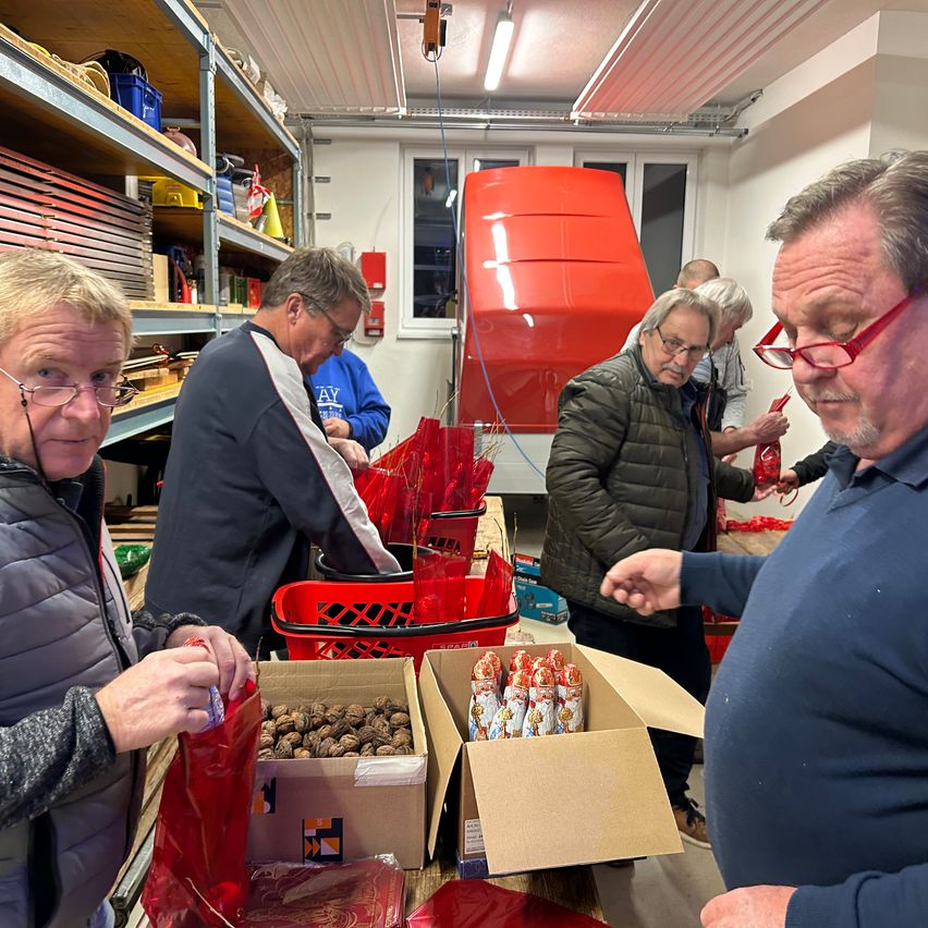 Eine Gruppe von Menschen in einem Raum, einige stecken Gegenstände in rote Tüten, andere betrachten Boxen mit Süßigkeiten und Walnüssen.