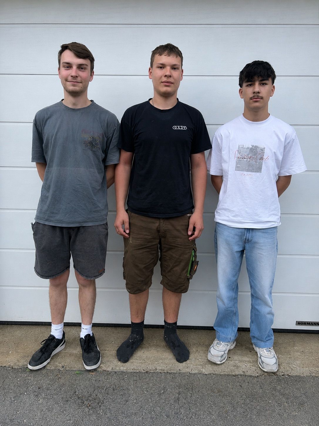 Three young men stand side by side, smiling. The left wears a grey t-shirt and shorts. The middle wears a black t-shirt with 'GADY' and brown shorts. The right wears a white t-shirt with a graphic and blue jeans.