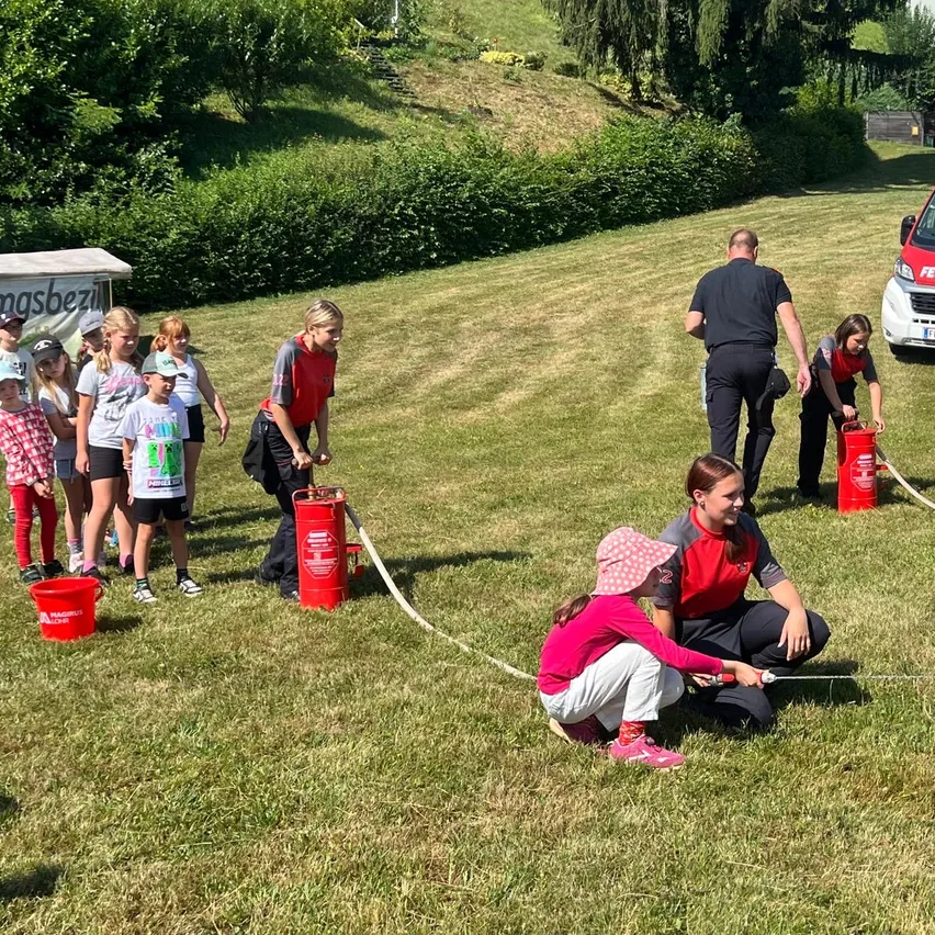 Eine Gruppe von Kindern und Erwachsenen versammelt sich auf einem Rasenfeld, einige halten rote Eimer und Schläuche, wahrscheinlich nehmen sie an einer wasserbezogenen Aktivität teil.