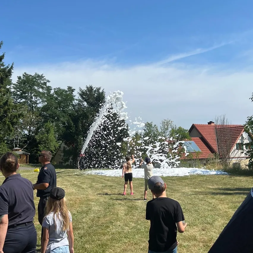 Eine Gruppe von Menschen, darunter Kinder, spielen an einem sonnigen Tag auf einer Wiese mit einem Wasserballon-Werfer.