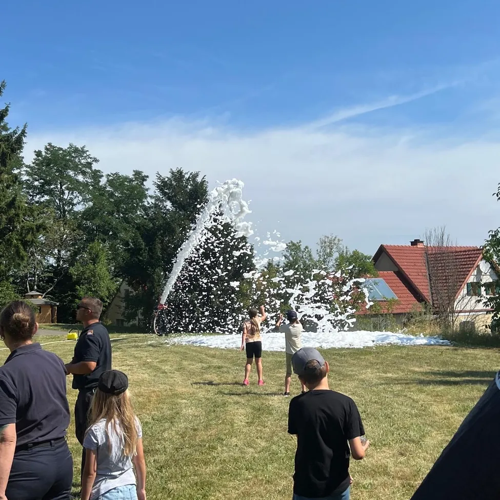 Eine Gruppe von Menschen, darunter Kinder, spielen an einem sonnigen Tag auf einer Wiese mit einem Wasserballon-Werfer.