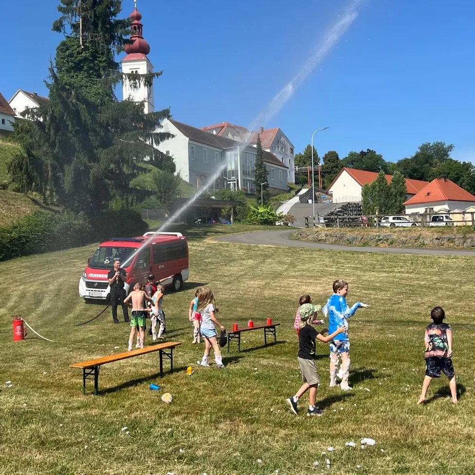 Eine Gruppe von Kindern spielt mit einer Wasserpistole in einem Park, während ein Feuerwehrauto in der Hintergrund Wasser sprüht.