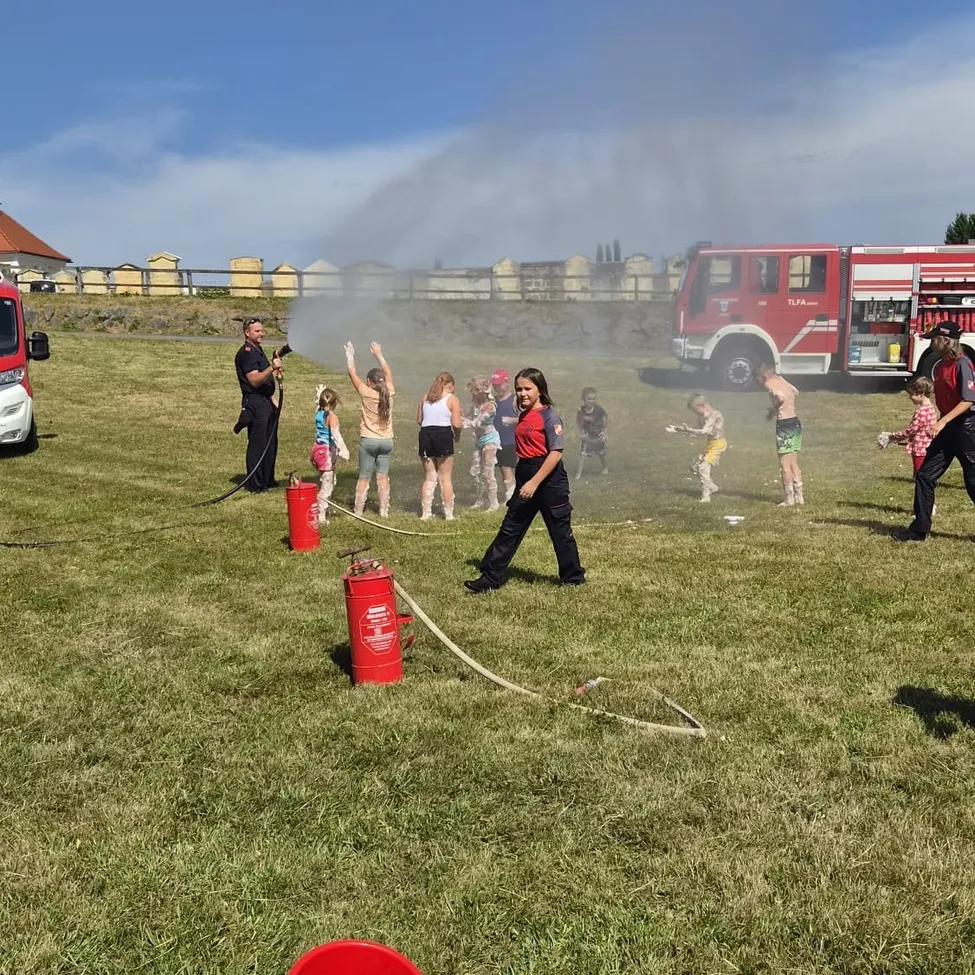 Eine Gruppe von Kindern und Erwachsenen spielt mit Wasser von einem Feuerwehrauto auf einem Rasen.