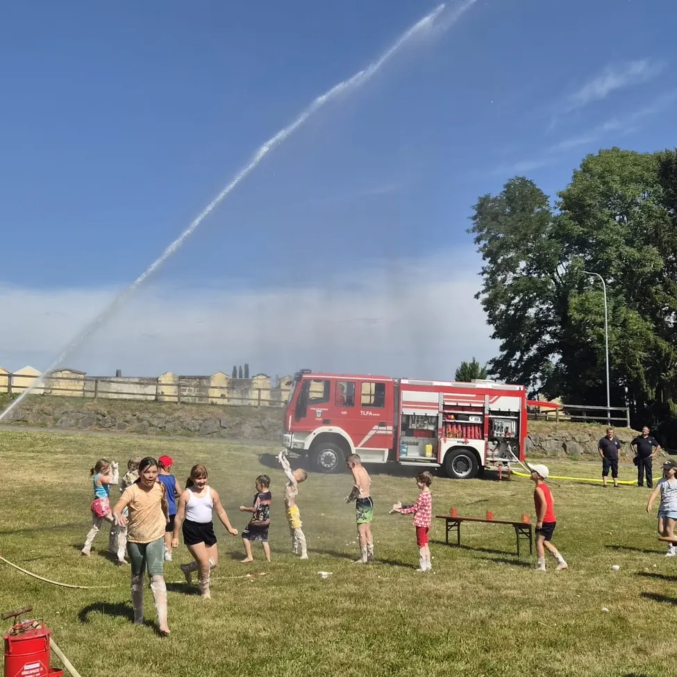 Eine Gruppe von Kindern spielt auf einem Feld, während ein Feuerwehrauto Wasser in die Luft sprüht.