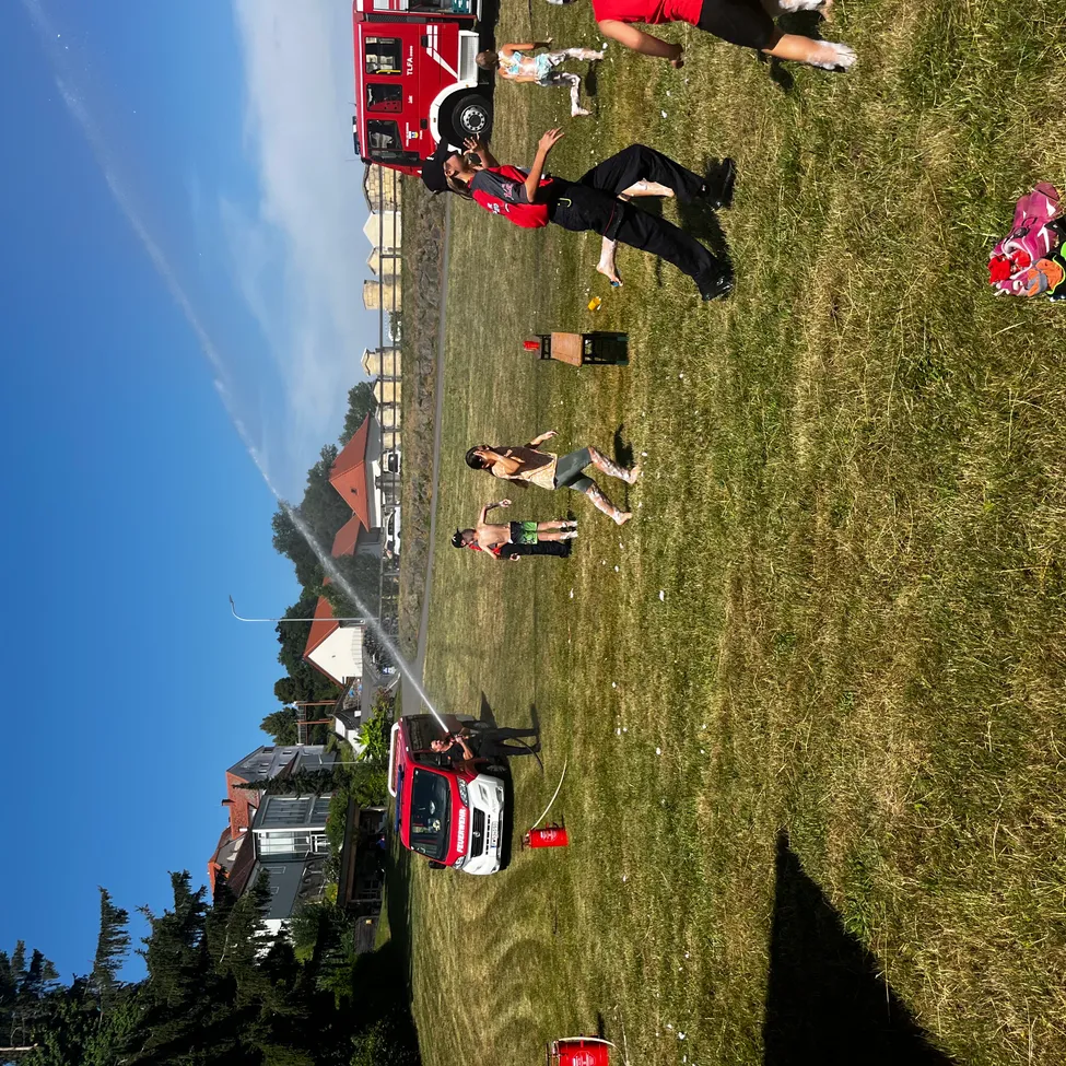 Eine Draufsicht auf eine Gruppe von Menschen, die auf einem Grasfeld spielen, mit einem Feuerwehrauto, das im Hintergrund Wasser sprüht.