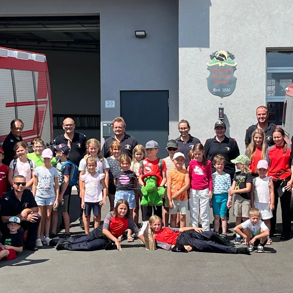 Eine Gruppe von Kindern und Erwachsenen posiert für ein Foto vor einer Feuerwache, wobei ein Feuerwehrauto im Hintergrund zu sehen ist.