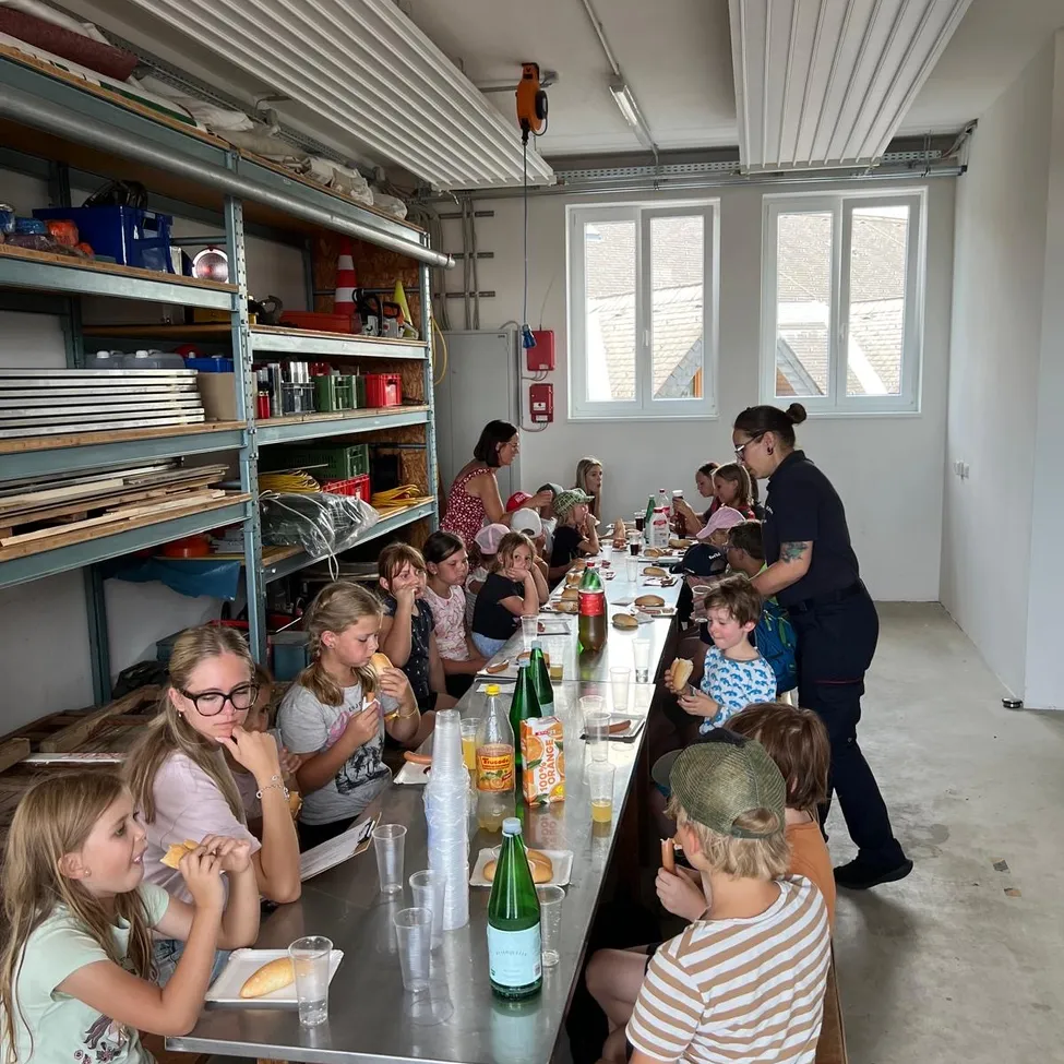 Eine Gruppe von Kindern und Erwachsenen sitzt an einem langen Tisch in einem Raum, isst Sandwiches und trinkt aus Bechern. Der Raum hat weiße Wände, Regale mit verschiedenen Gegenständen und Fenster. Eine Frau in einem schwarzen Shirt steht am Ende des Tisches.