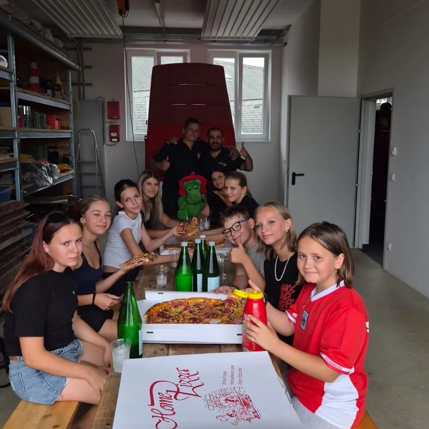Eine Gruppe von Menschen, einschließlich Kindern, sitzt um einen Tisch mit Pizzaschachteln und Flaschen, lächelt und posiert für ein Foto in einem Raum.