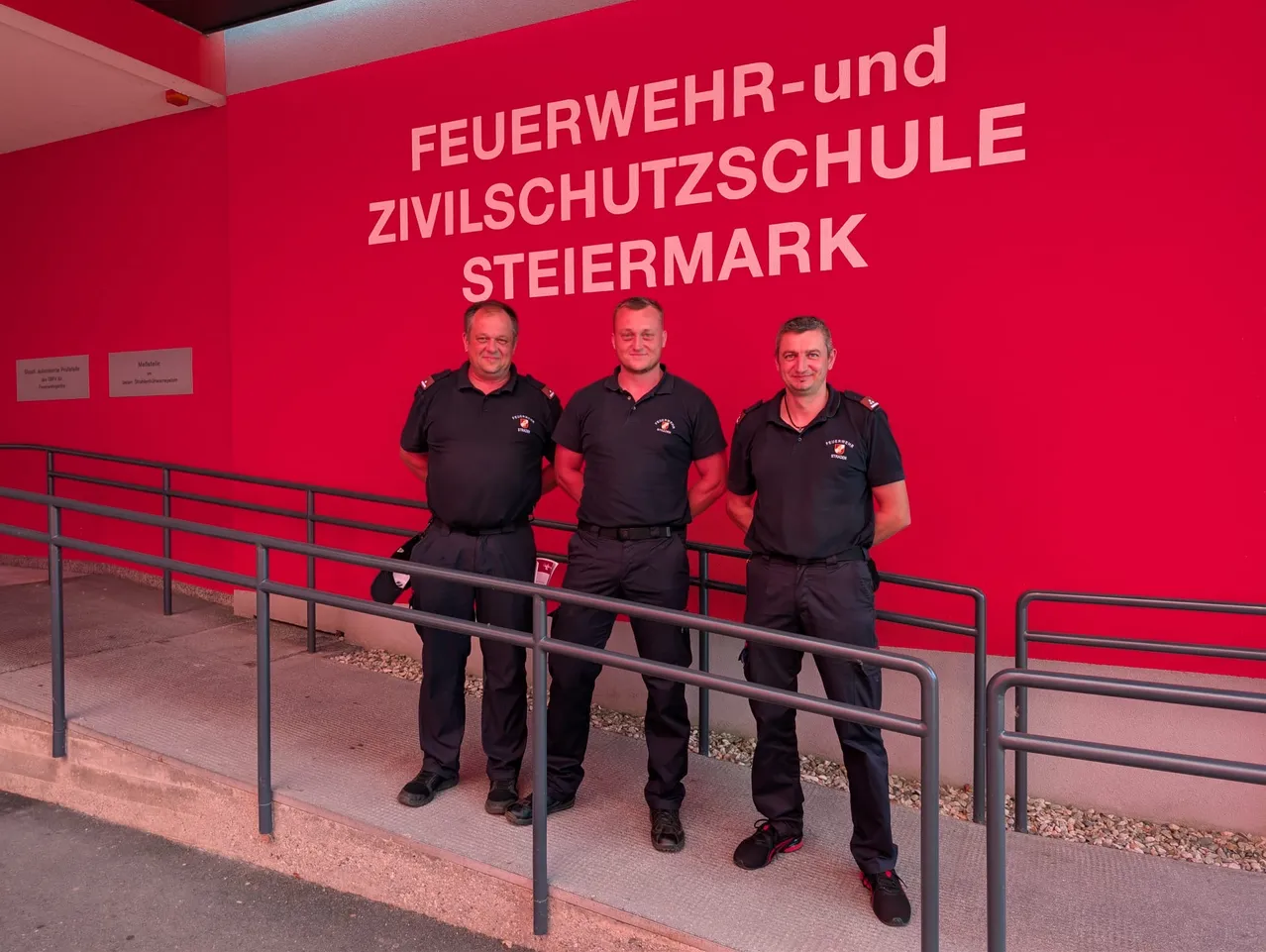 Drei Männer in Uniform stehen vor einer roten Wand mit weißem Text, der 'Feuerwehr-und Zivilschutzschule Steiermark' lautet.