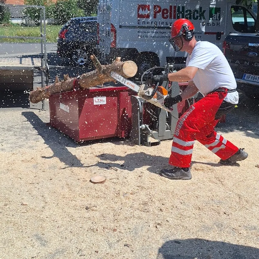 Ein Mann in Helm und Sicherheitsausrüstung säg ein Stück Holz mit einer Kettensäge im Freien. Ein weißer Lieferwagen und ein schwarzes Auto sind im Hintergrund geparkt.