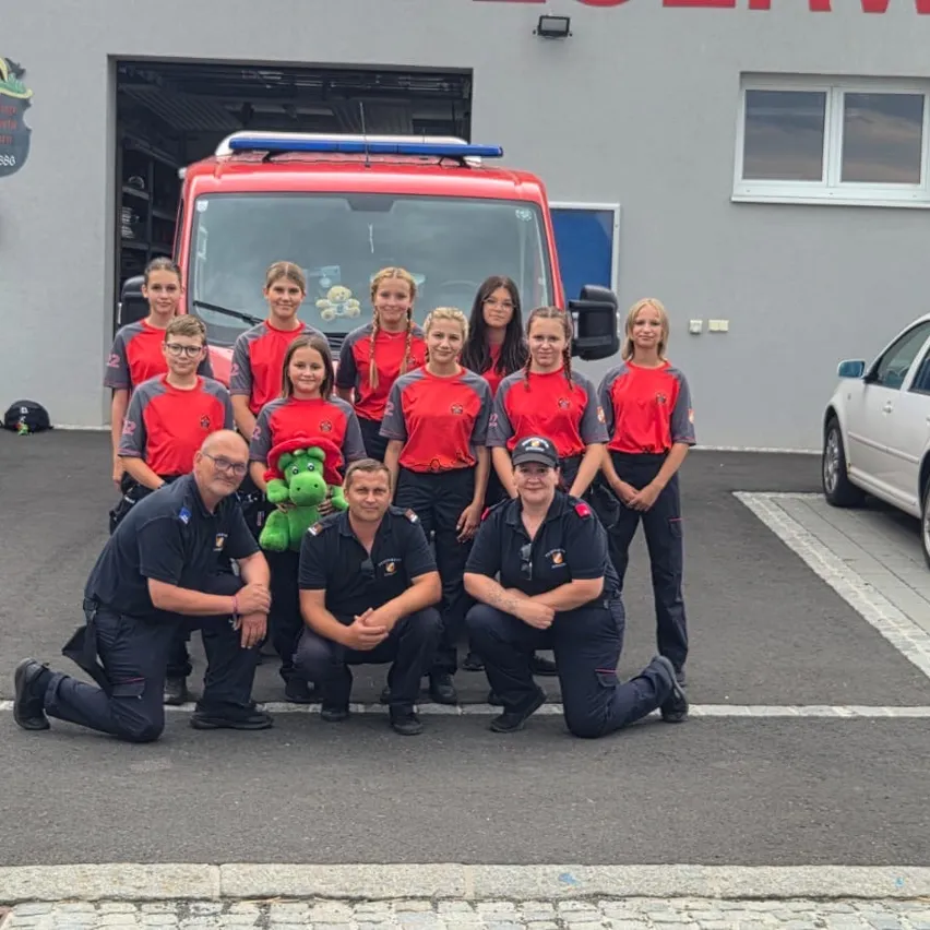 Eine Gruppe Feuerwehrleute in roten Uniformen posiert für ein Foto vor einem Feuerwehrwagen. Zwei Männer knien, einer mit einem grünen Kuscheltier. Ein Gebäude mit Garage und Auto steht hinter ihnen.