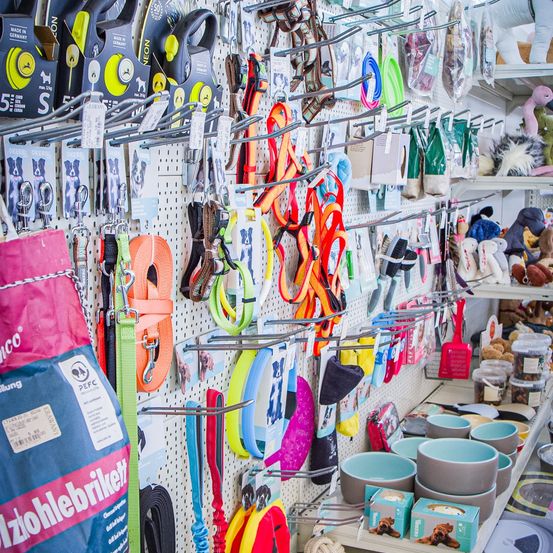 Ein Laden mit verschiedenen Haustierprodukten ausgestellt. Mehrere Leinen, Halsbänder und Geschirre hängen an einem Lochbrett. Schüsseln, Spielzeug und Hundefutter sind auf Regalen. Eine große Tasche mit dem Wort 'Training' ist sichtbar.