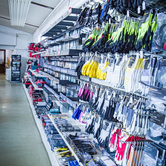 Ein Gang in einem Geschäft mit Regalen, die mit verschiedenen Produkten gefüllt sind. Die Regale sind mit einer Vielzahl von Handschuhen, Werkzeugen und anderen Artikeln bestückt. Der Boden ist gefliest und die Wände sind weiß.