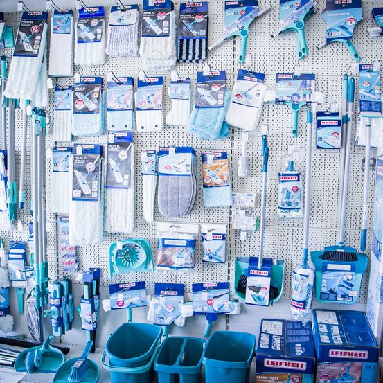 An einer Wand sind verschiedene Reinigungsprodukte zu sehen, darunter Besen, Staubwedel und Mop. Die Produkte sind in Reihen angeordnet, mit sichtbaren Etiketten und Logos auf der Verpackung. Die Wand hat ein weißes und blaues Farbschema.