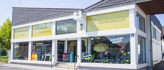 Fassade eines Geschäfts namens Krawany mit zwei Schildern, eines sagt Baustoffe & Bedachung und das andere Ofenkollektion. Im Schaufenster sind verschiedene Artikel wie Regenschirme und Spielzeug zu sehen.
