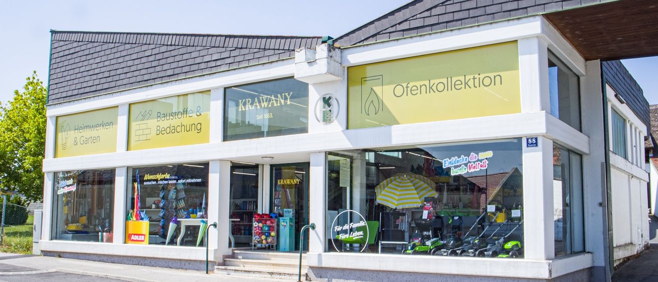 Fassade eines Geschäfts namens Krawany mit zwei Schildern, eines sagt Baustoffe & Bedachung und das andere Ofenkollektion. Im Schaufenster sind verschiedene Artikel wie Regenschirme und Spielzeug zu sehen.