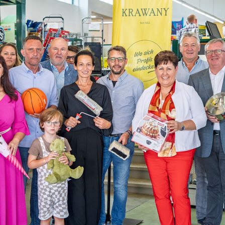 Eine Gruppe von Erwachsenen und Kindern posiert bei einer Veranstaltung. Eine Frau hält eine Box und eine Dose Farbe, während eine andere Frau einen Korb hält. Ein Kind hält einen pelzigen Dinosaurier. Sie stehen neben einem Schild mit der Aufschrift 'KRAWANY'.