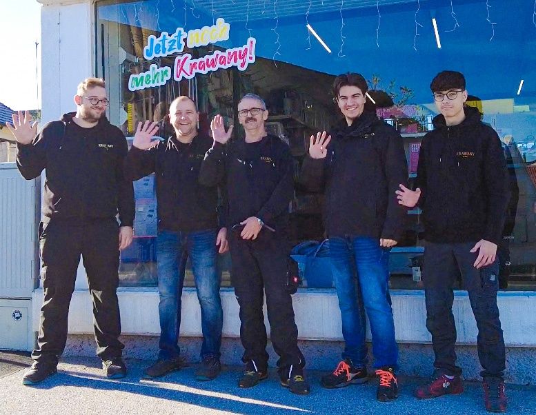 Fünf Männer in schwarzen Jacken posieren für ein Foto vor einem Geschäft mit einem blauen Banner. Sie stehen eng beieinander, lächeln und machen Handgesten.
