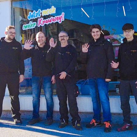 Fünf Männer in schwarzen Jacken posieren für ein Foto vor einem Geschäft mit einem blauen Banner. Sie stehen eng beieinander, lächeln und machen Handgesten.