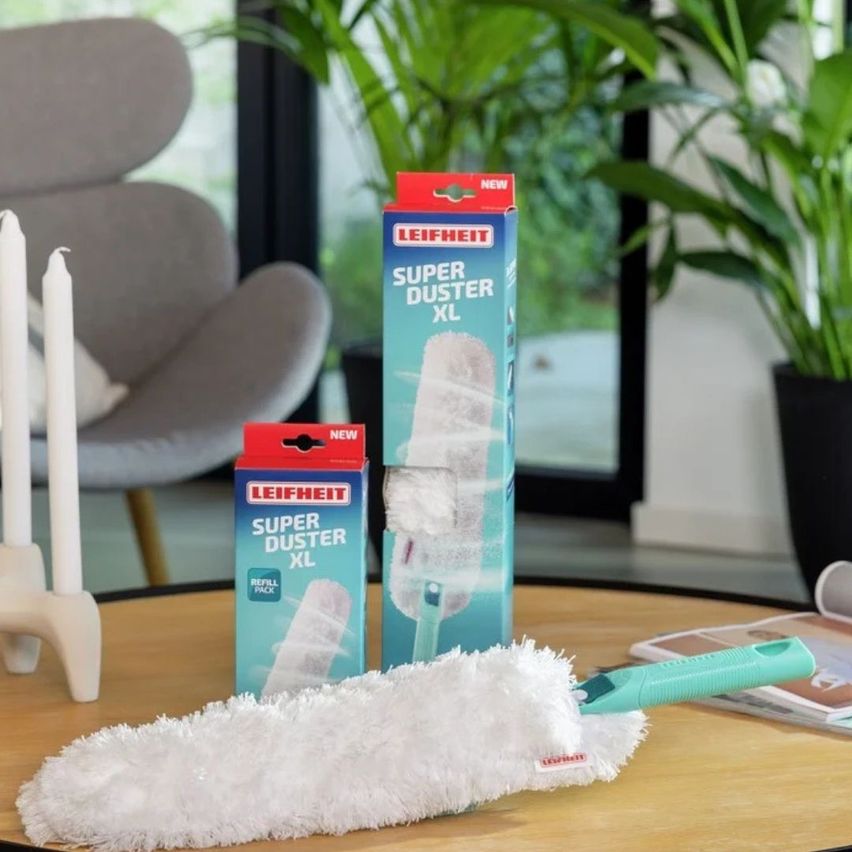 Ein Tisch zeigt einen Leifheit Super Duster XL Staubwedel mit seiner Nachfüllpackung und Box auf einer Holzoberfläche, mit einem grauen Stuhl und Kerzen im Hintergrund.