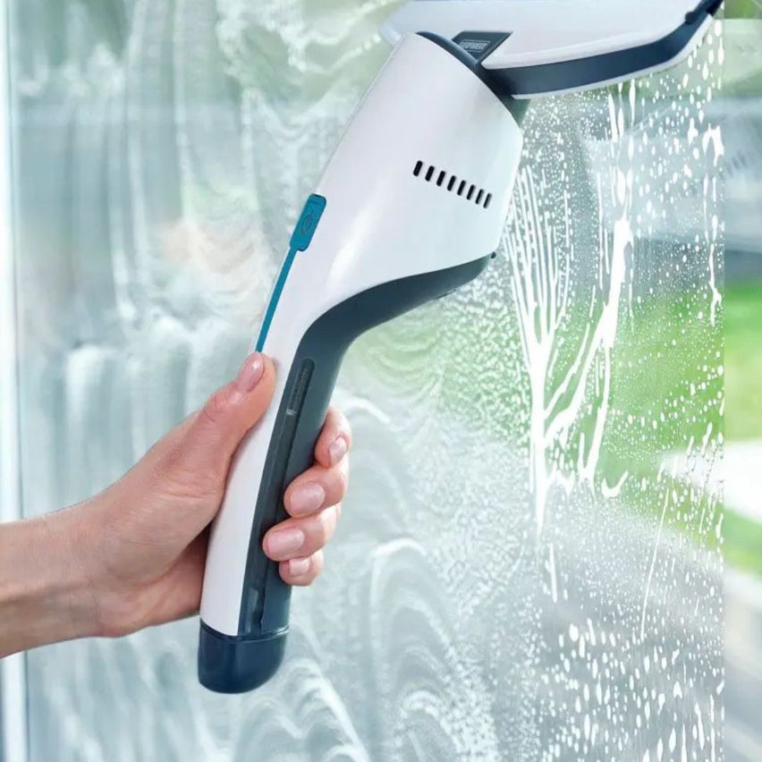 Eine Person benutzt ein weißes und blaues Handreinigungsgerät, um ein Glasfenster zu reinigen und Wassertropfen auf die Oberfläche zu sprühen.