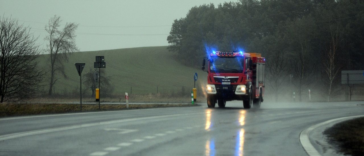 Ein roter Feuerwehrwagen fährt mit blauen Lichtern bei Regen auf einer nassen Straße. Bäume und ein grasbedeckter Hügel sind im Hintergrund zu sehen.