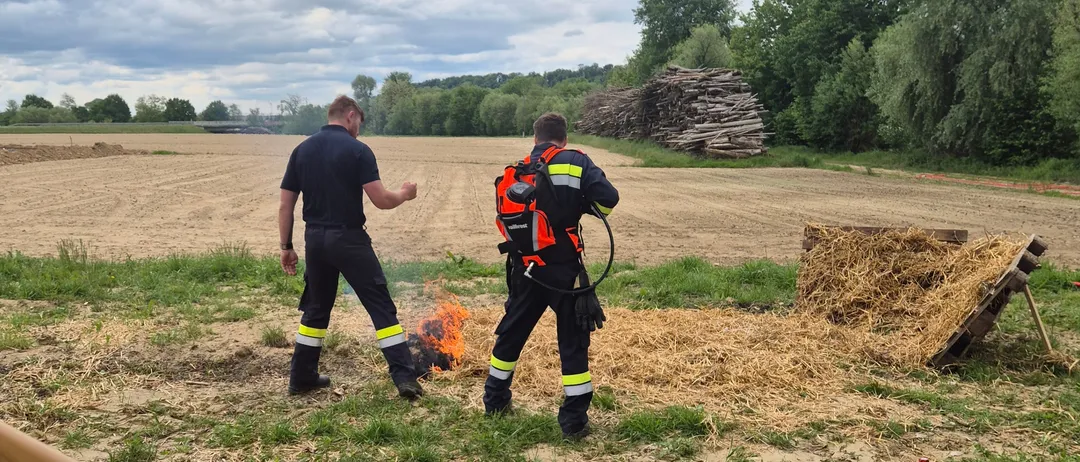 Zwei Feuerwehrleute in Schutzkleidung stehen in einem offenen Feld neben einem kleinen Feuer am Boden, mit einem Holzstock und einem blauen Gaszylinder in der Nähe.