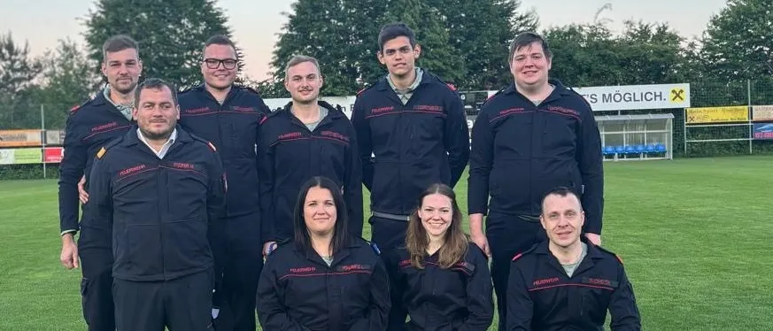 Eine Gruppe von Menschen in Uniformen posiert für ein Foto auf einem Feld mit Helmen und Trophäen vor ihnen. Dahinter sind Bäume und ein Gebäude mit einem Schild.