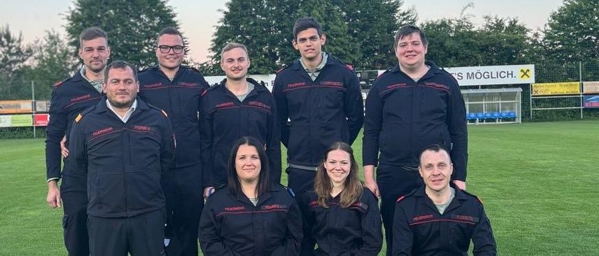 Eine Gruppe von Menschen in Uniformen posiert für ein Foto auf einem Feld mit Helmen und Trophäen vor ihnen. Dahinter sind Bäume und ein Gebäude mit einem Schild.