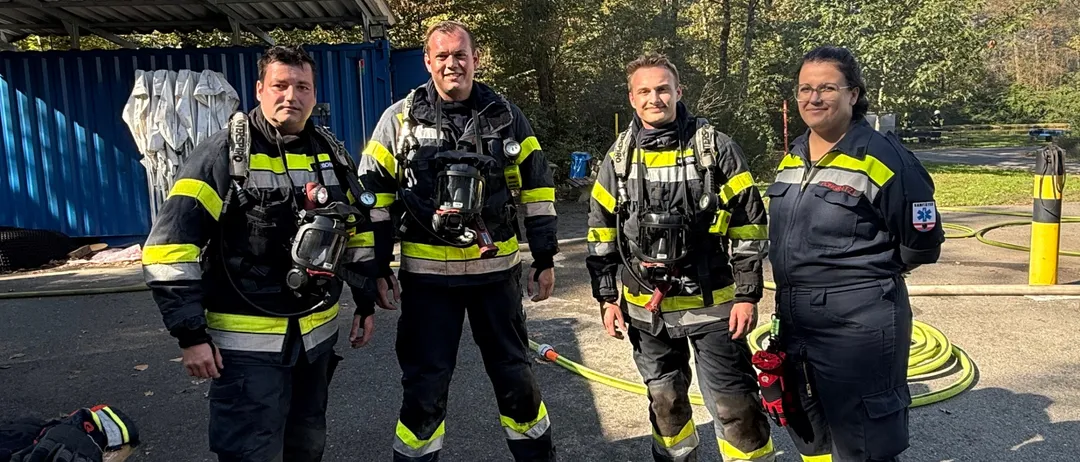 Vier Feuerwehrleute in voller Ausrüstung stehen auf einem Parkplatz. Dahinter eine blaue Struktur mit Bäumen und einem Feuerhydranten.