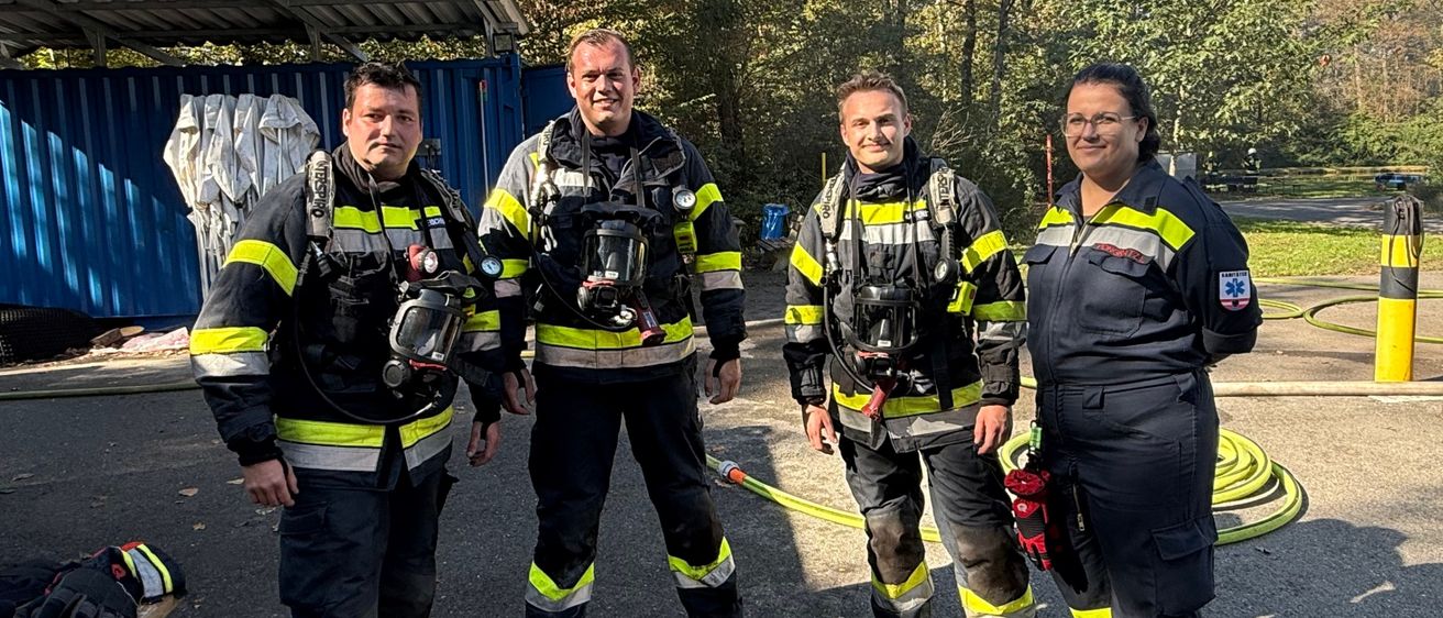 Vier Feuerwehrleute in voller Ausrüstung stehen auf einem Parkplatz. Dahinter eine blaue Struktur mit Bäumen und einem Feuerhydranten.