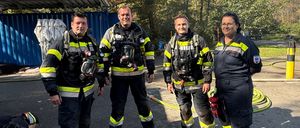 Vier Feuerwehrleute in voller Ausrüstung stehen auf einem Parkplatz. Dahinter eine blaue Struktur mit Bäumen und einem Feuerhydranten.