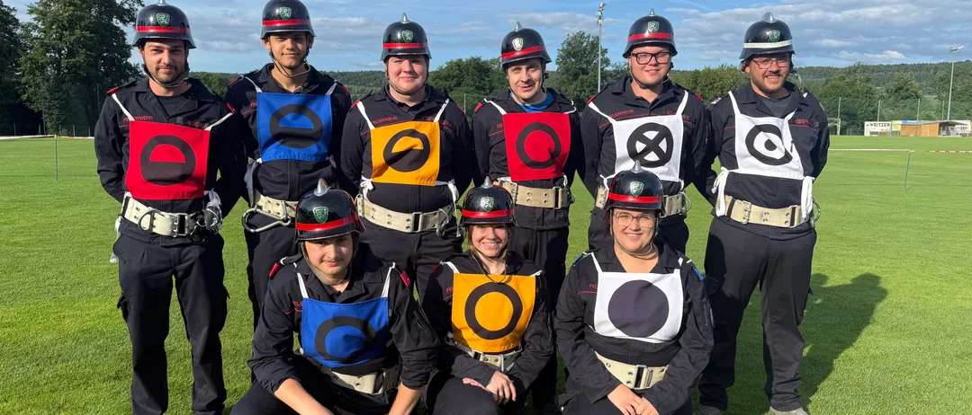 Eine Gruppe von Menschen in Uniformen, Helmen und Sicherheitswesten mit verschiedenen Symbolen posiert für ein Foto auf einem Rasenfeld.