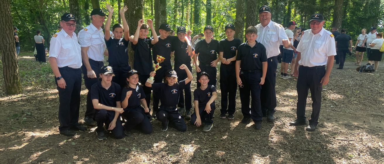 Eine Gruppe von Menschen in Uniform posiert für ein Foto im Wald, einige halten eine Trophäe. Das Gebiet ist von hohen Bäumen umgeben.