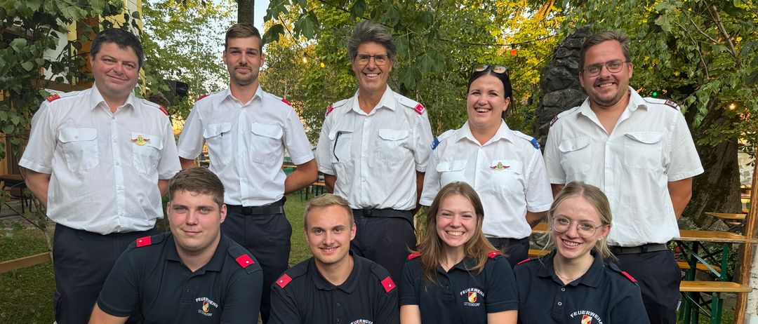 Eine Gruppe von Feuerwehrleuten in Uniform posiert für ein Foto in einem Park mit Bäumen im Hintergrund.