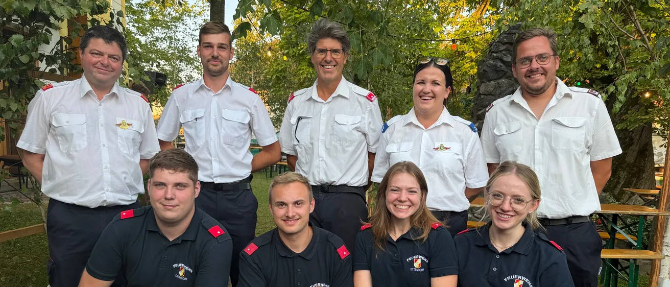 Eine Gruppe von Feuerwehrleuten in Uniform posiert für ein Foto in einem Park mit Bäumen im Hintergrund.