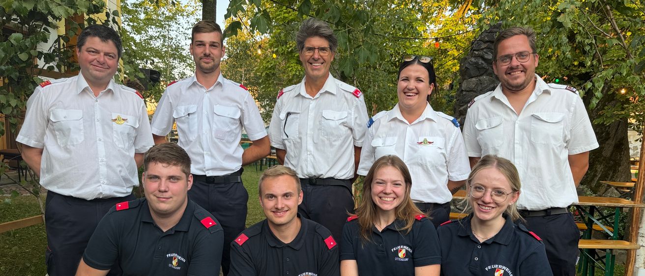 Eine Gruppe von Feuerwehrleuten in Uniform posiert für ein Foto in einem Park mit Bäumen im Hintergrund.