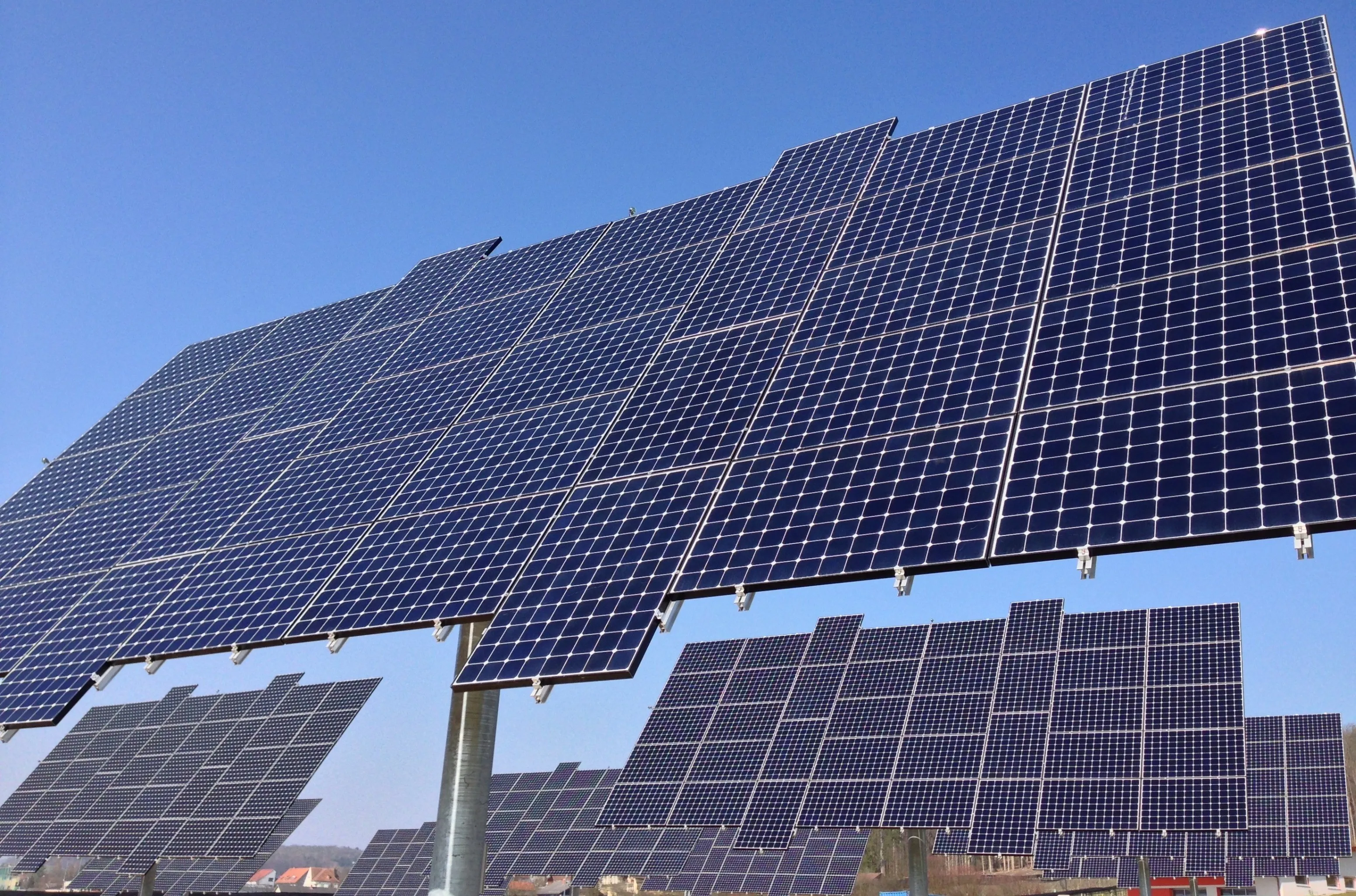 Eine große Anordnung von Solarpanelen, die nach oben gerichtet sind, unter einem klaren blauen Himmel mit mehreren Gebäuden im Hintergrund sichtbar.