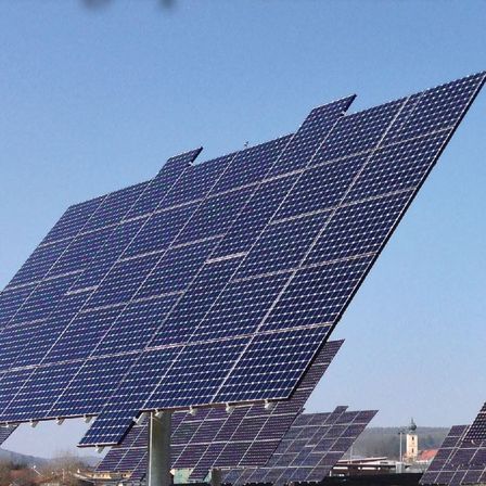 Solarmodule sind in Reihen unter einem klaren blauen Himmel angeordnet. Die Module sind nach oben gerichtet und fangen Sonnenlicht ein. Der Text 'tmannsdorf Erneuerbare duktion GmbH & Co KG' ist oben in weiß geschrieben.