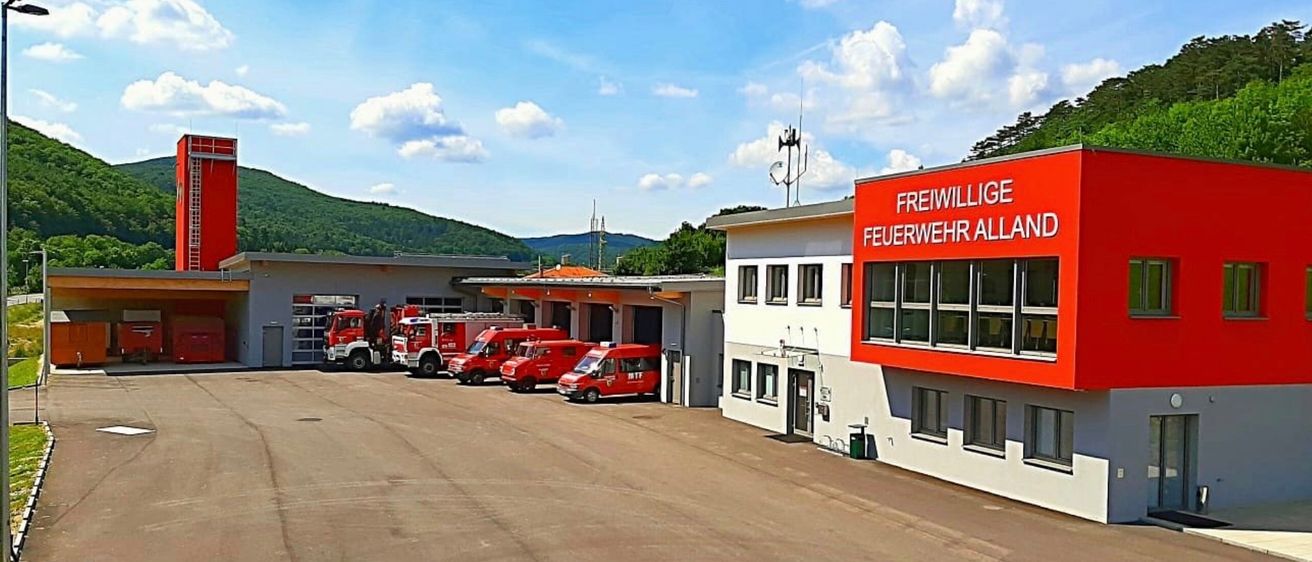 Eine Feuerwehrstation mit mehreren roten Lkws, die an einem sonnigen Tag vor dem Gebäude geparkt sind, mit Bergen im Hintergrund.