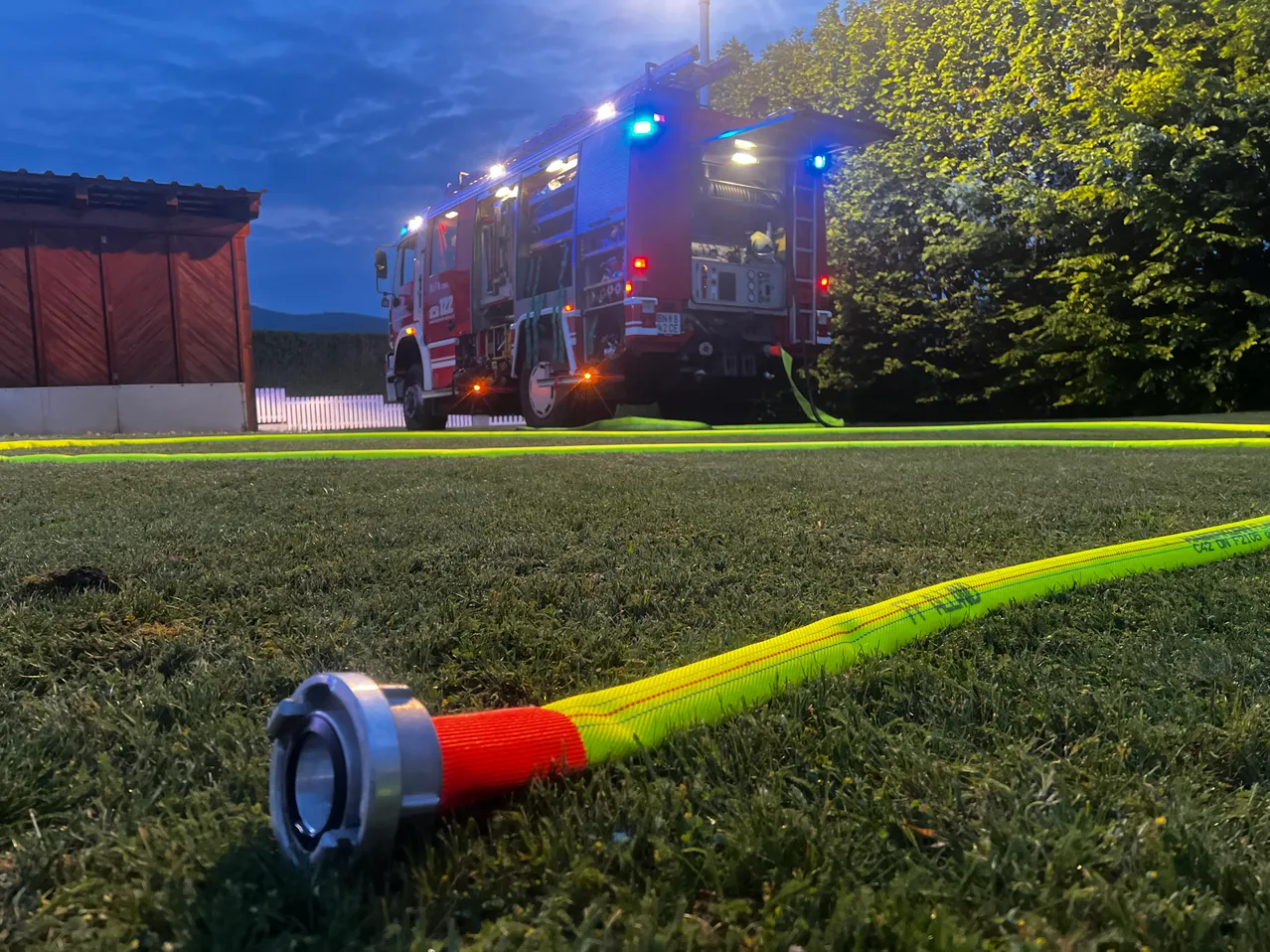 Ein roter Feuerwehrwagen mit blauen und roten Lichtern steht bei Dämmerung auf einem grünen Rasen, mit einem gelben Schlauch auf dem Boden.
