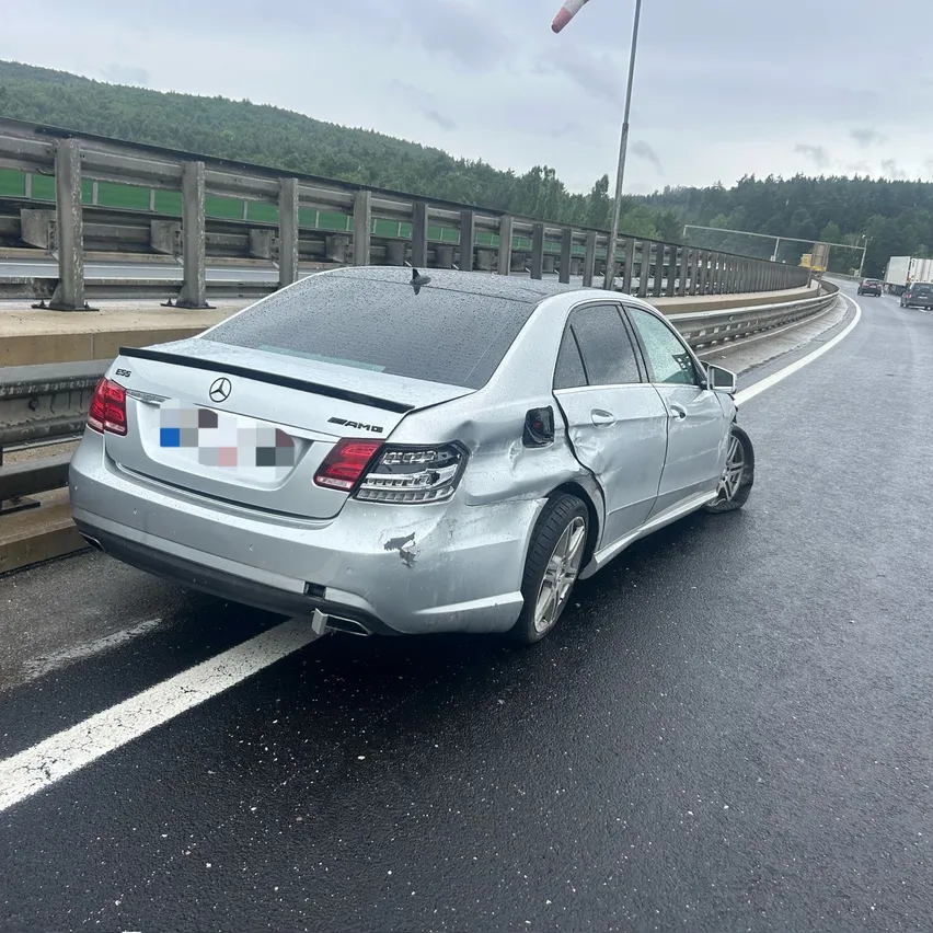 Ein silberner Mercedes-Benz E-Klasse ist auf der Seite einer Autobahn geparkt, hinten beschädigt. Es steht in der Nähe einer Leitplanke, mit Hügeln und Bäumen im Hintergrund.