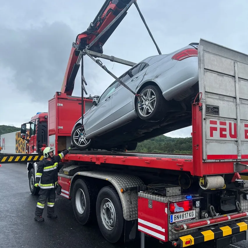 Ein Feuerwehrmann in Uniform sichert ein silbernes Auto auf einem Flachbett-Lkw mit einem Kran. Das Auto ist auf dem Kopf mit den Rädern nach oben. Der Lkw hat einen rot-weißen Körper mit dem Wort 'FEU' an der Seite.